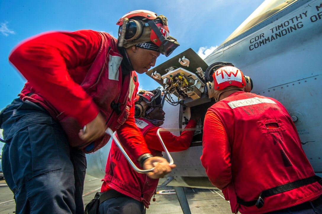 Three sailors, wearing red, use tools to work on an aircraft panel.
