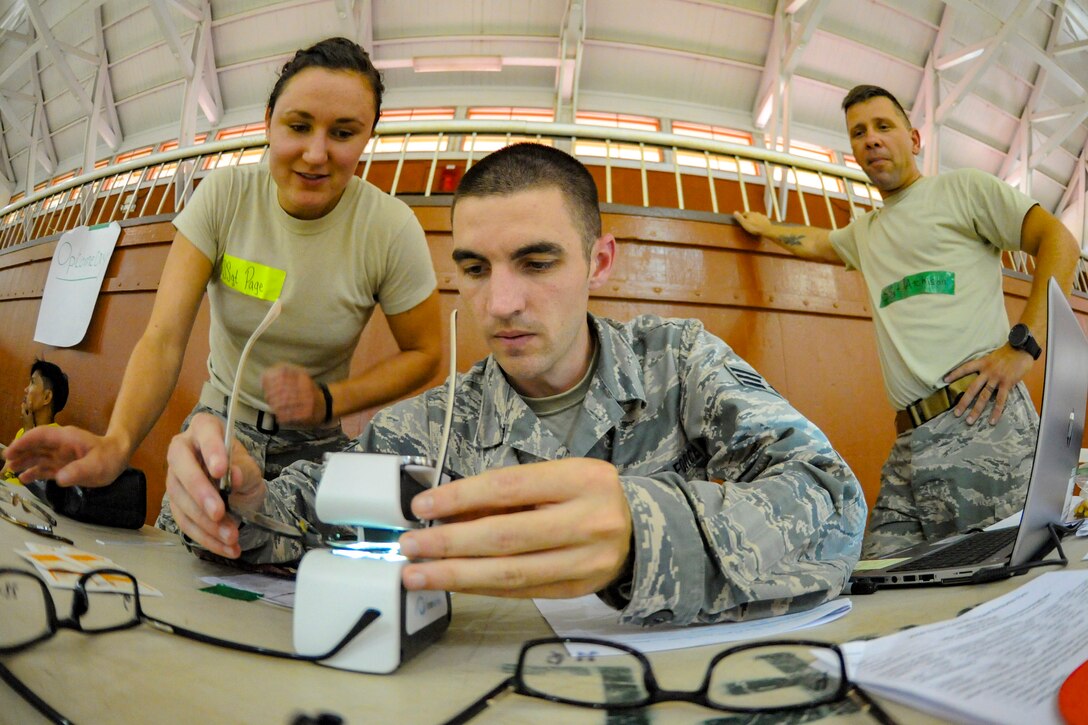 Two airman use a device to examine eyeglasses on a table, while another airman watches.