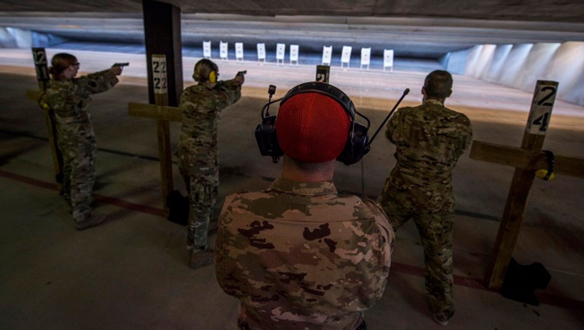 Airmen at a gun range shoot at targets with pistols as an instructor watches them form behind