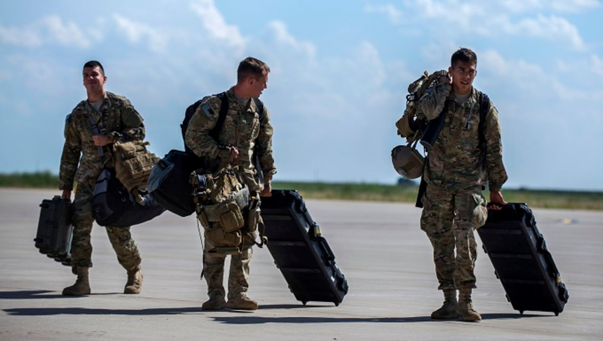 Three Airmen walk along the flight line with suitcases and packed bags