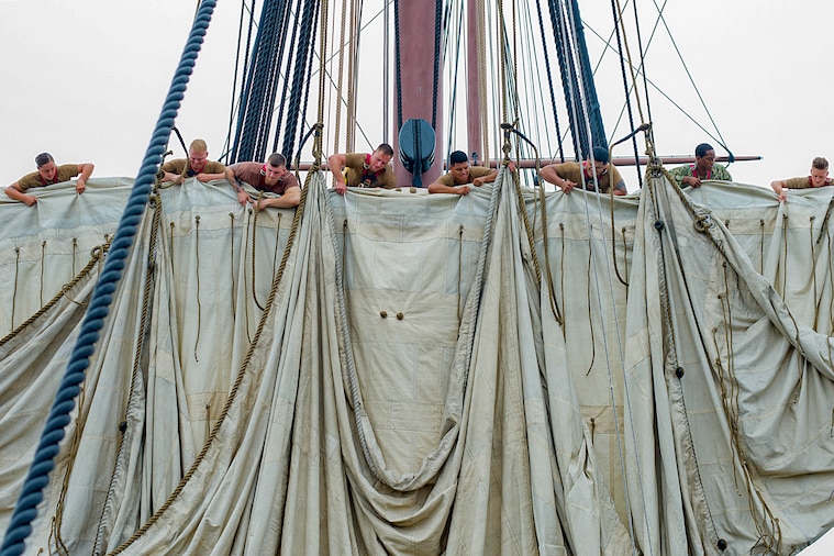 Sailors unfurl a sail high above the deck of a ship.