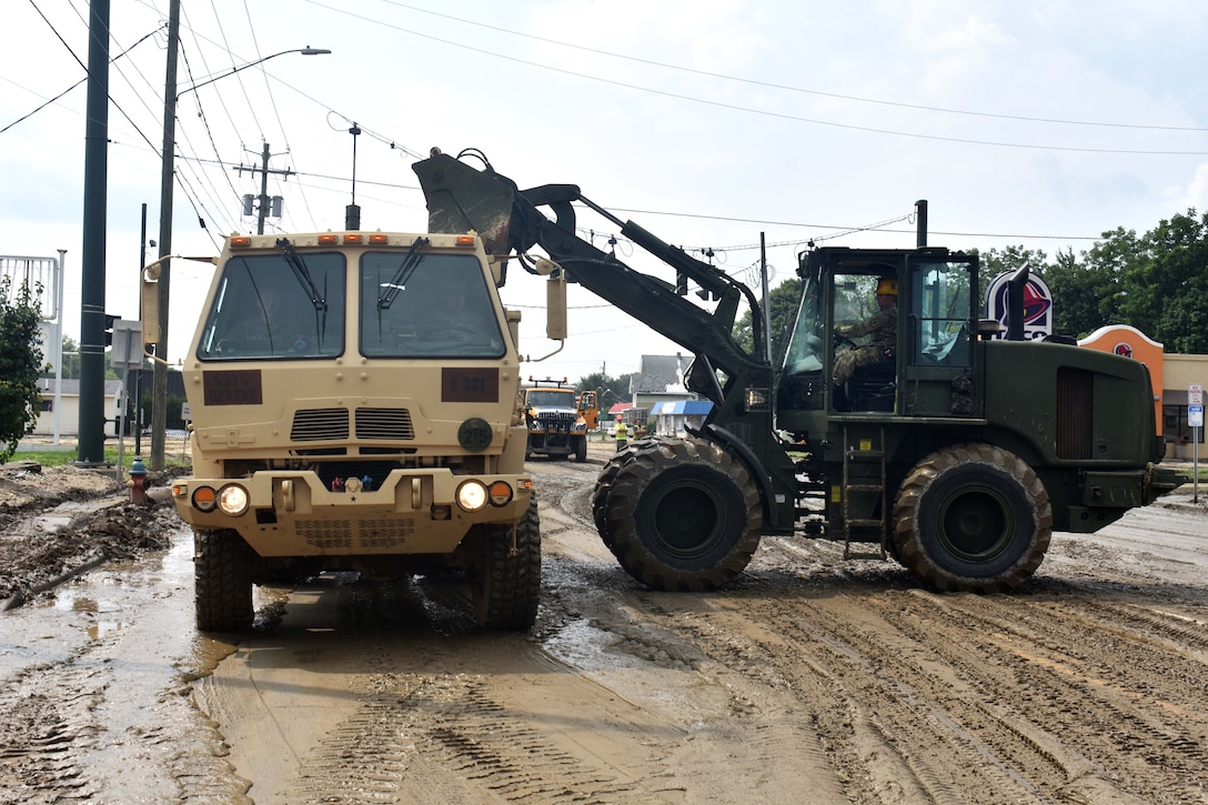 A soldier operates a bulldozer to clear mud and debris from streets.
