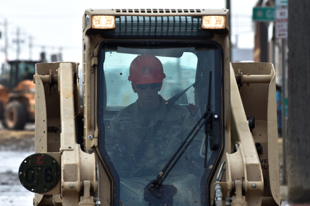 A soldier operates a bobcat bulldozer to clear mud and debris from streets.