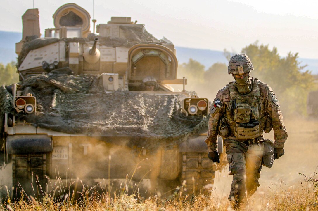 A soldier walks in front of a tank through a field.