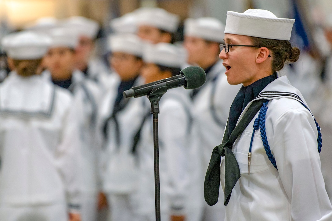 Sailors listen as another sailor speaks into a microphone.