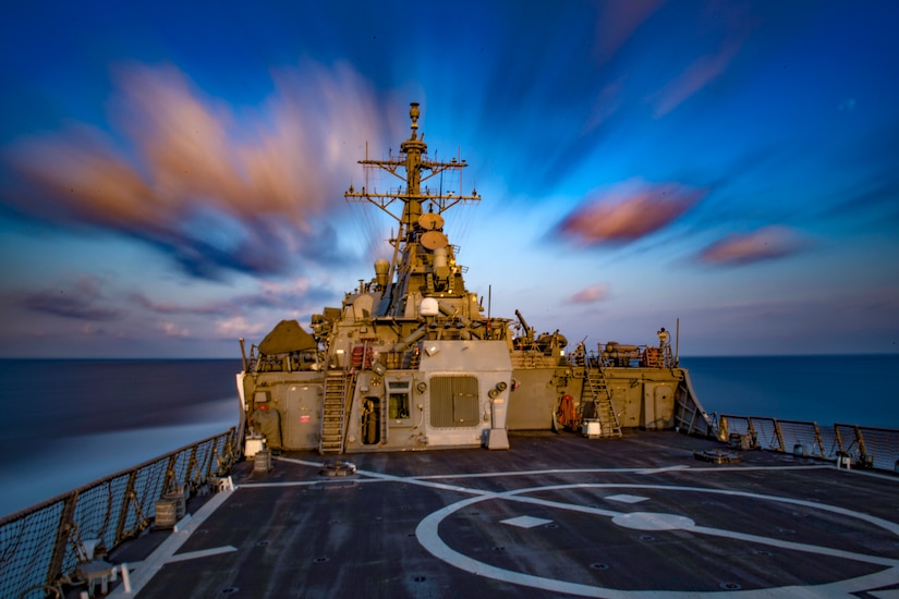 A navy ship passes under the cloudy sky.