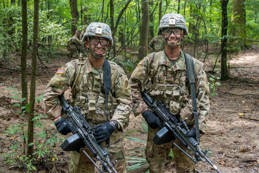 Two soldiers wearing camouflage paint stand in a forest.