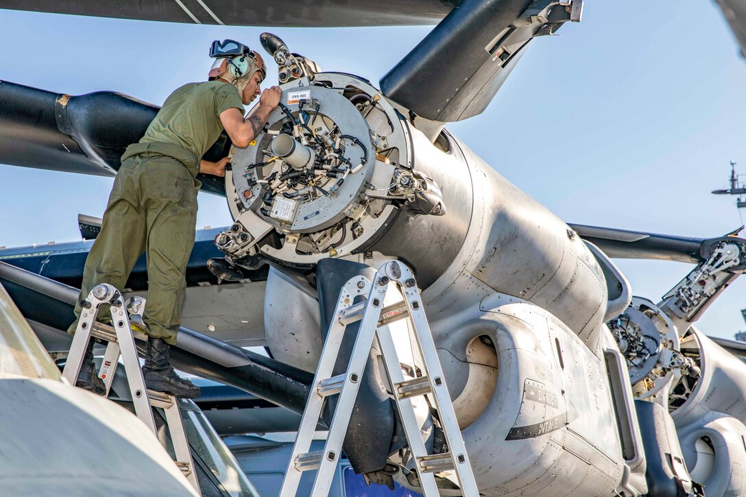 A Marine on a ladder uses a handheld tool on an aircraft.