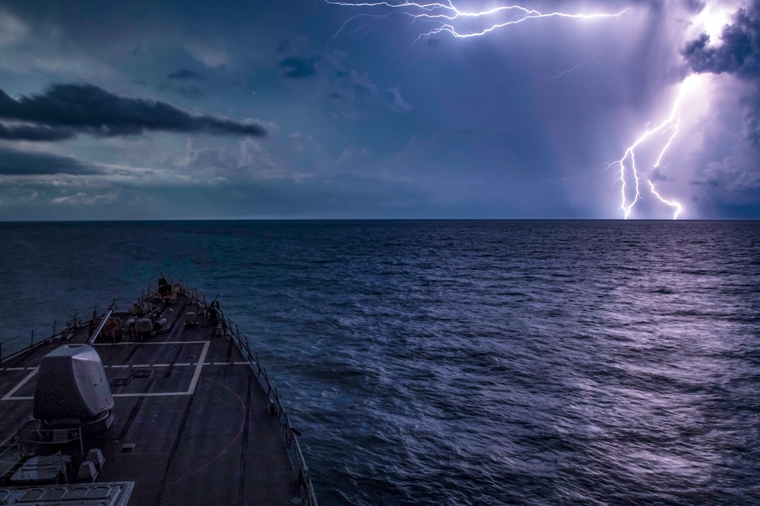 A ship travels in the sea as lightning in the distance illuminates a purplish sky.