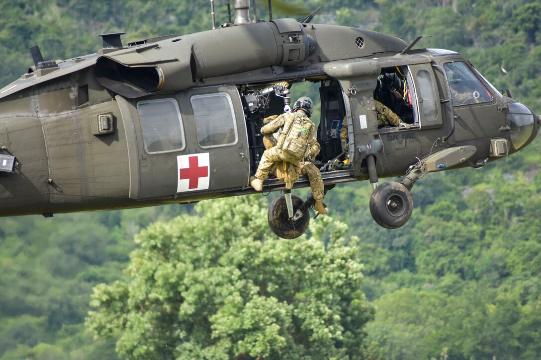 A soldier hangs out the open door of a hovering helicopter.