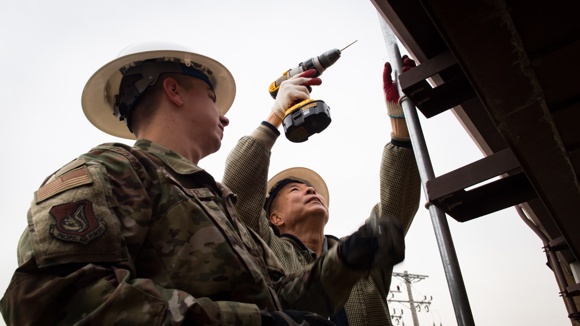 U.S. Air Force Airman 1st Class Jesse Slater, left, and Yong Ui Song, fire alarm technicians with the 51st Civil Engineer Squadron, drill holes into the side of a metal rooftop to secure a new antenna on Osan Air Base, Republic of Korea, Nov. 29, 2018.