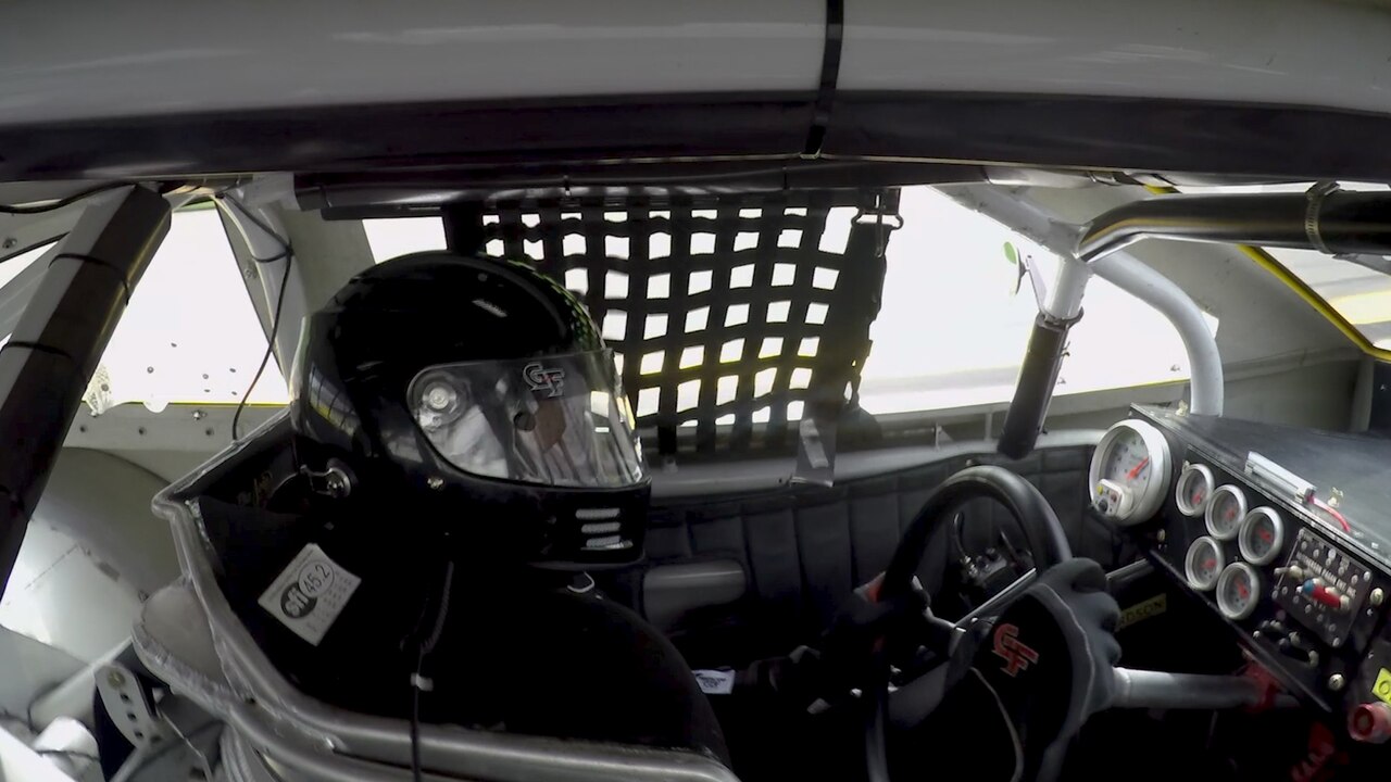 A helmeted race car driver sits behind the wheel of a race car.