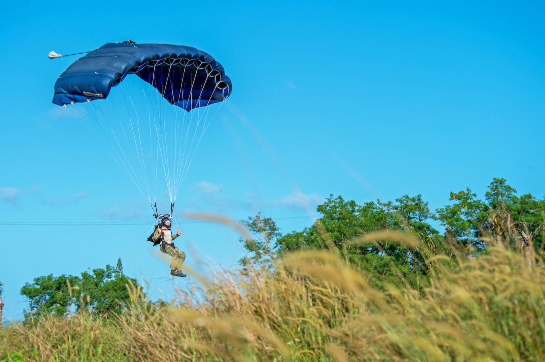 A sailor steers a parachute while coming in for a landing among shrubs.