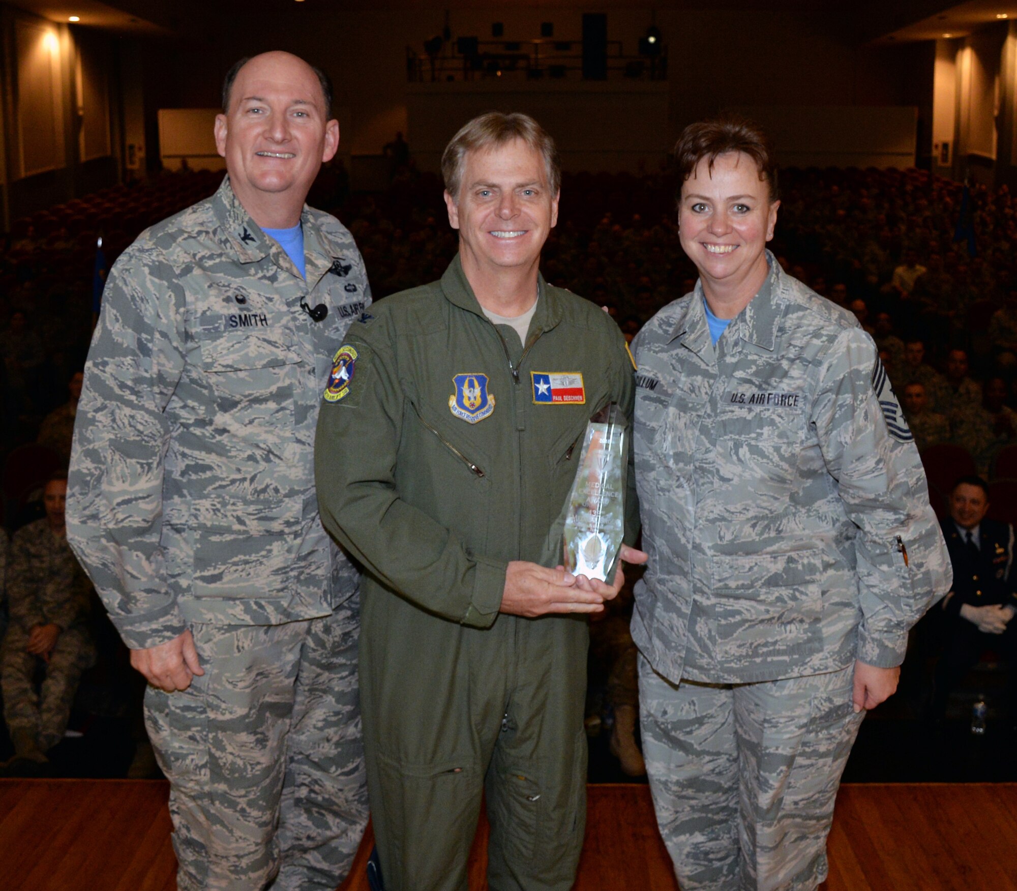 433rd Airlift Wing Commander Thomas K. Smith, Jr. and 433rd AW Command Chief Master Sgt. Shana Cullum stand with Col. Paul Deschner, 433rd Aeropace Medicine Squadron commander, who accepts the Raincross Trophy on behalf of the 433rd Medical Group, during commander's call Dec. 2, 2018 in the Bob Hope theater at Joint Base San Antonio-Lackland, Texas.
