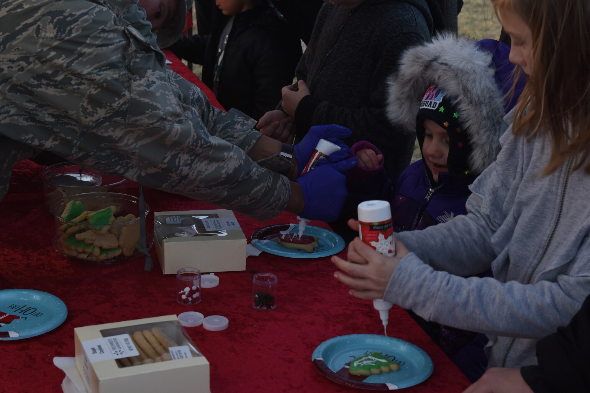 A volunteer helps children decorate cookies during the Tree Lighting Ceremony at the parade field on Goodfellow Air Force Base, Texas, Dec. 4, 2018. The children were able to take the decorations and cookies home with them or eat the cookies with some hot chocolate offered at the event. (U.S. Air Force photo by Airman 1st Class Zachary Chapman/Released)
