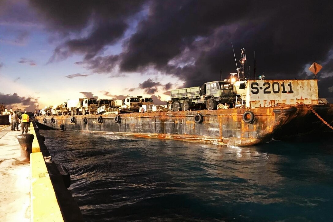 A barge carrying military tactical vehicles and equipment departs a pier.