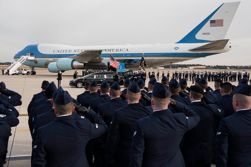 A group of service members salute as a hearse pulls alongside Air Force One.