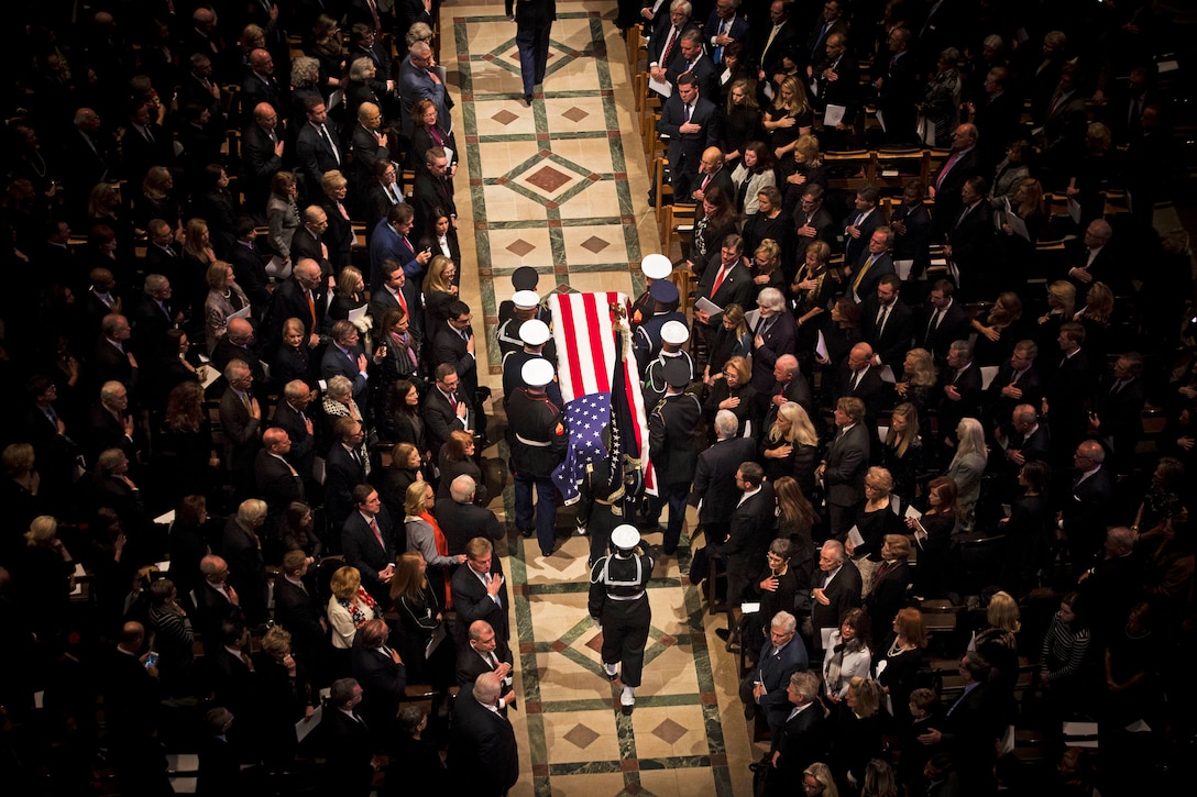 Seen from overhead,  service members carry a casket down the aisle while funeral attendees look on.