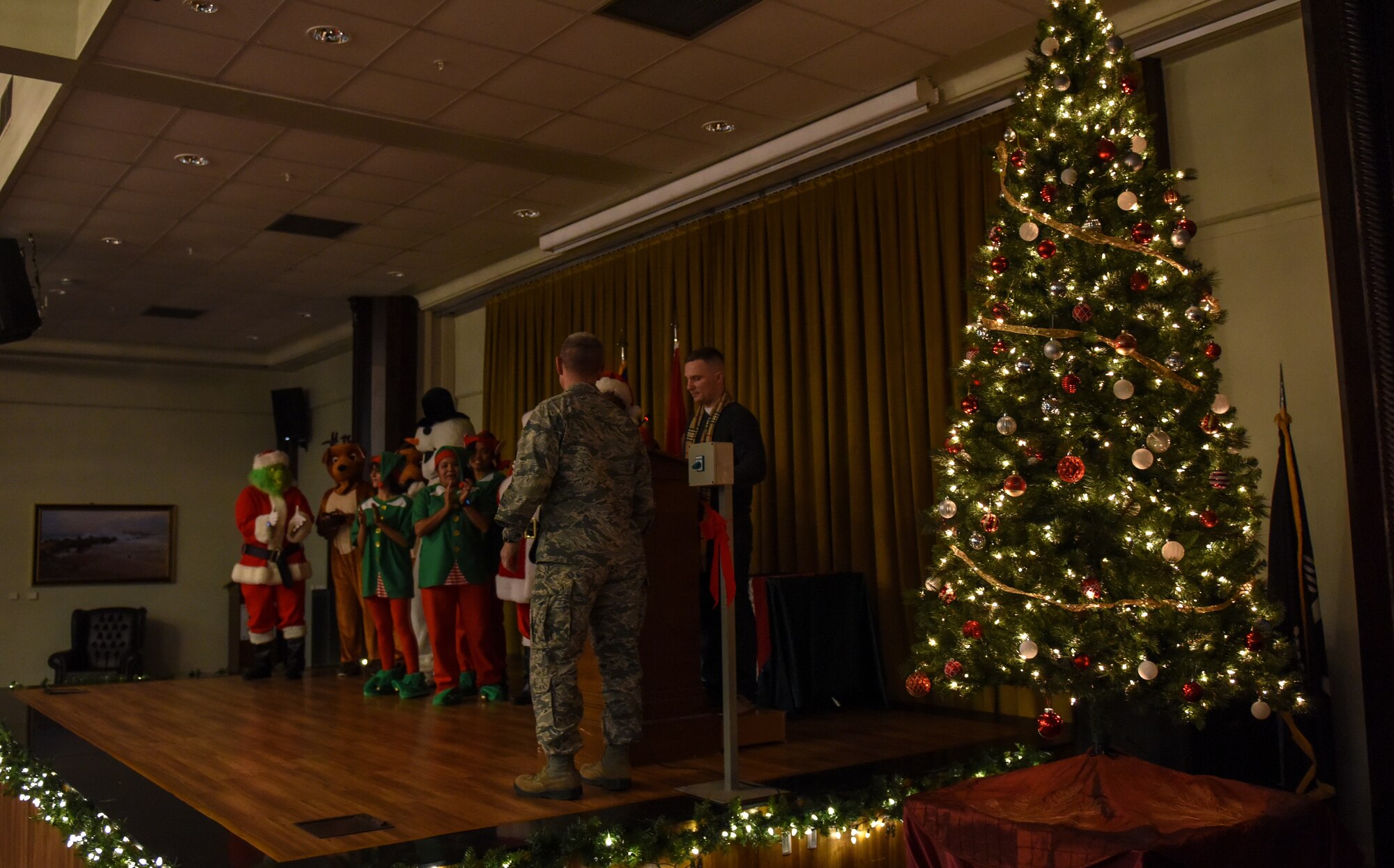 Airmen dressed as holiday characters join U.S. Air Force Col. Britt Hurst on stage during a holiday tree lighting ceremony.