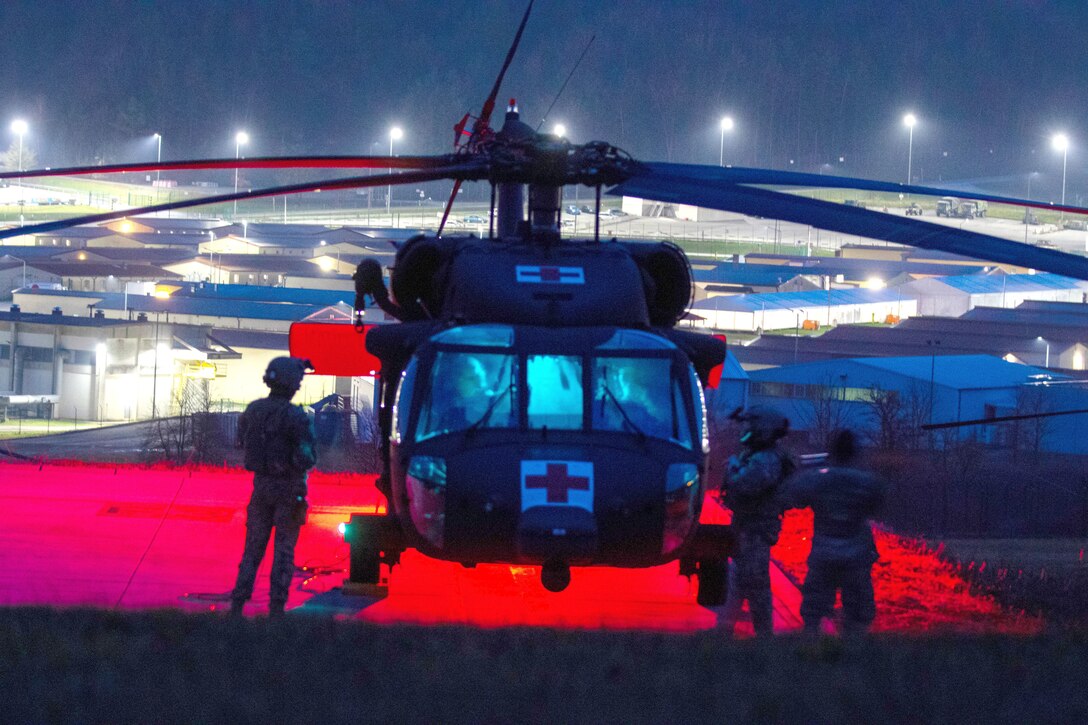 Soldiers stand around a helicopter at night with bright lights behind them.