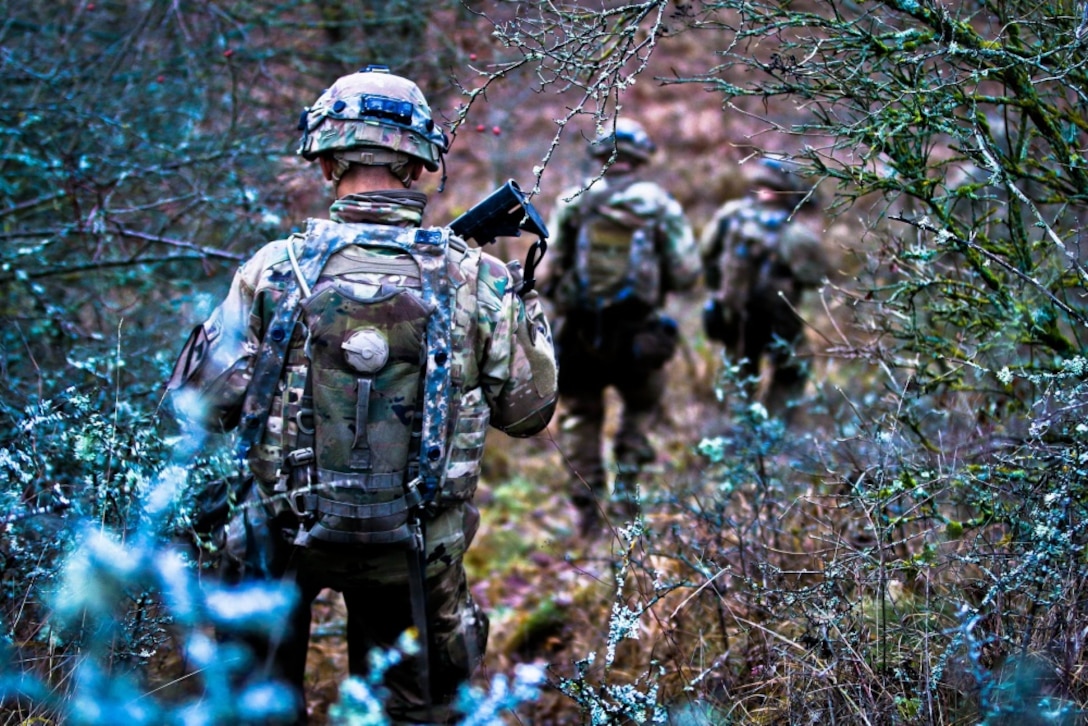 Soldiers walk through brush in a valley.