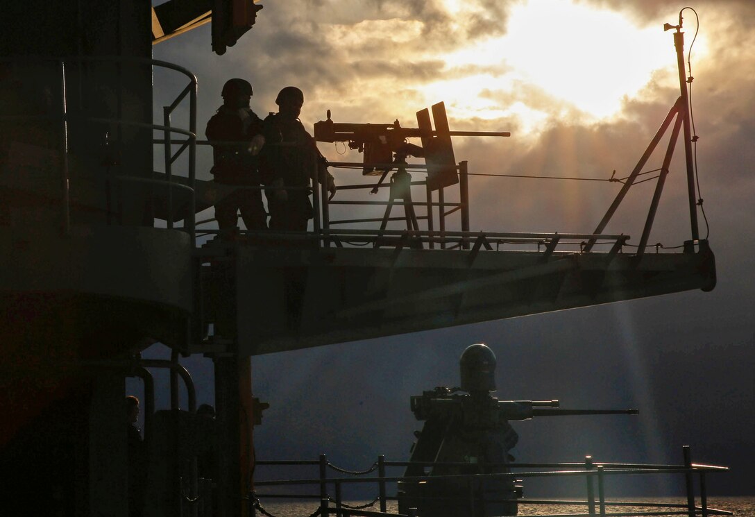 Two sailors stand watch near a gun mount on the upper deck of a ship.