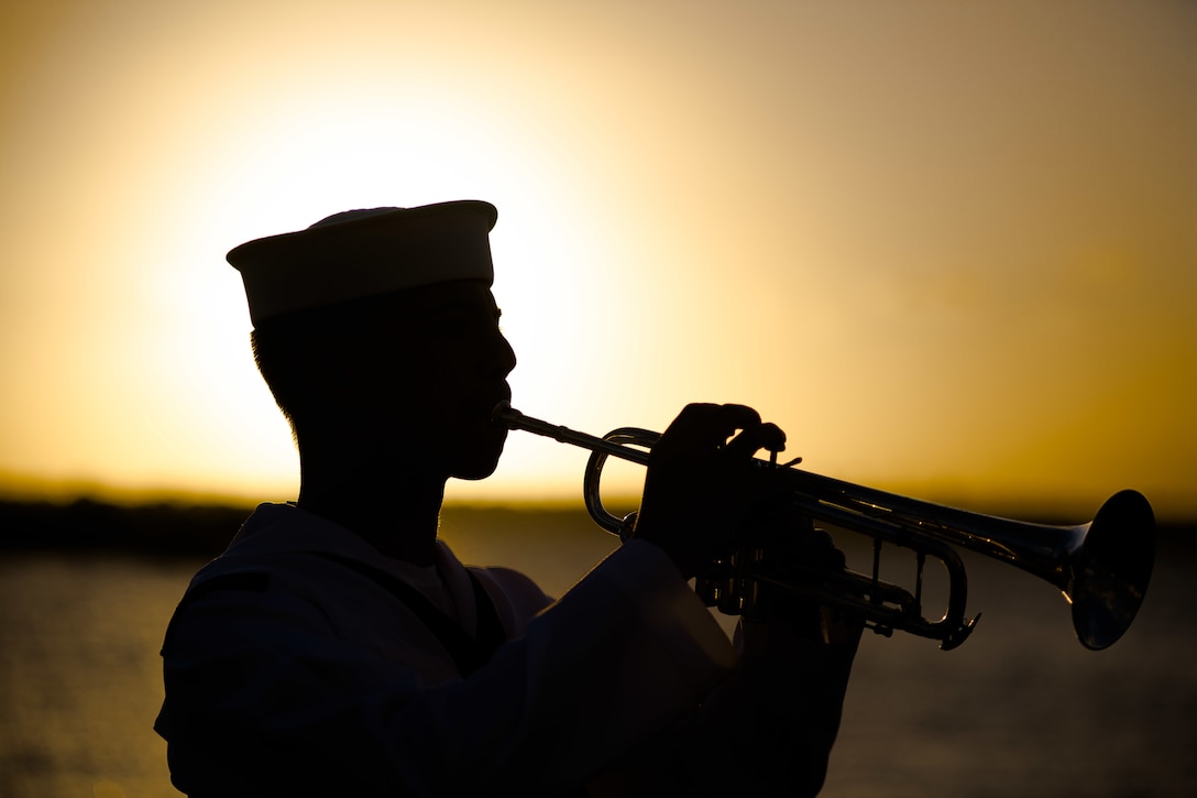 A sailor plays taps on a trumpet at sunset.