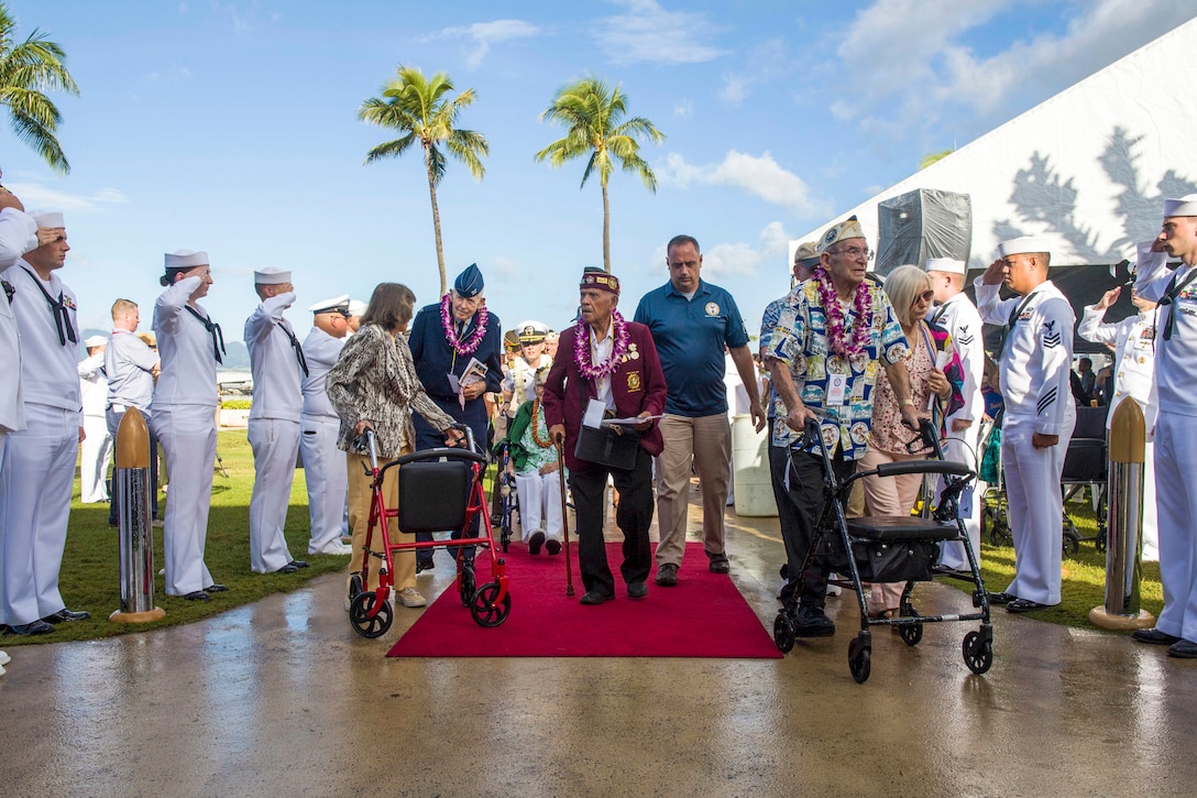 Sailors salute World War II veterans walking on a red carpet in front of palm trees.
