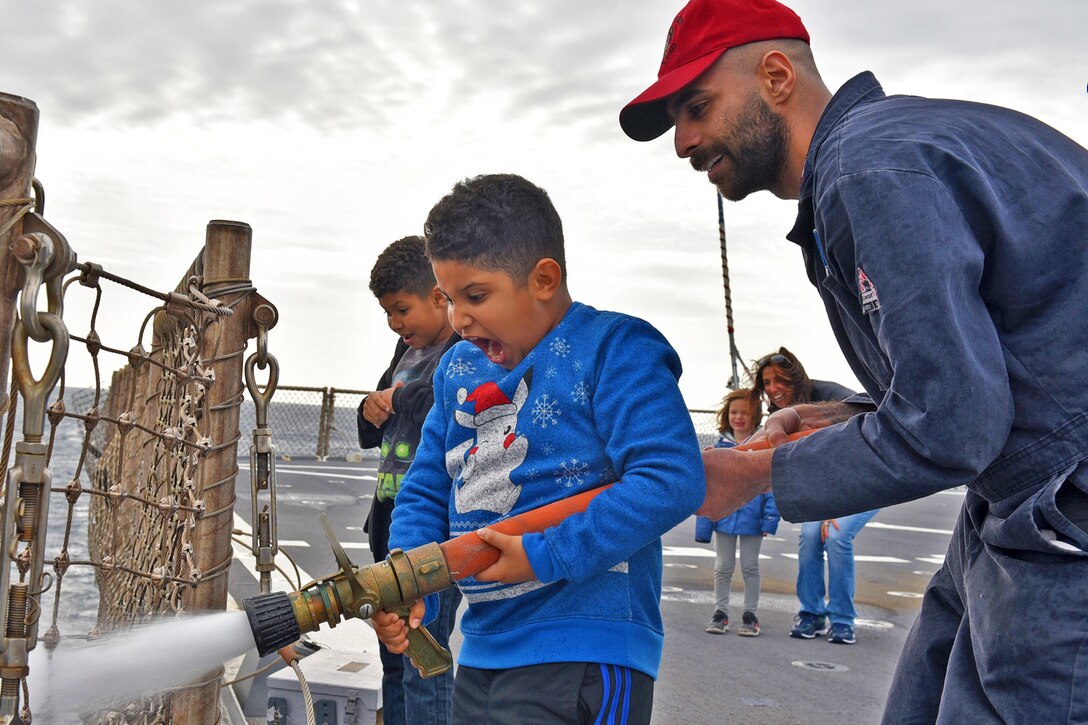 A sailor helps a child hold a fire hose on a ship.