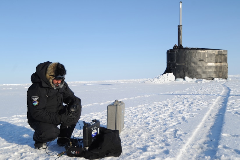 A sailor kneels on ice in front of a submarine.