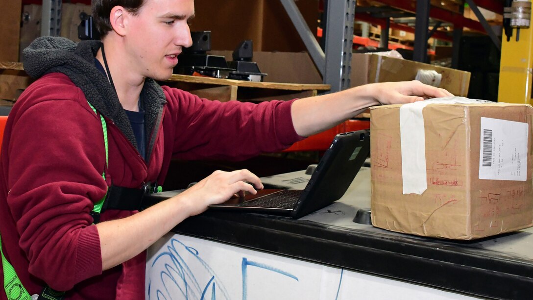 A DLA Distribution employee uses a tablet computer to research information from a cardboard box in front of him.