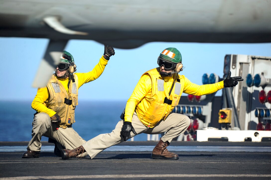 Two kneeling sailors in yellow tops point to launch an aircraft on a flight deck.