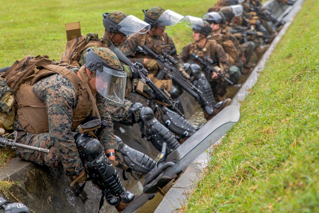A line of Marines wearing riot gear stand behind a barrier.