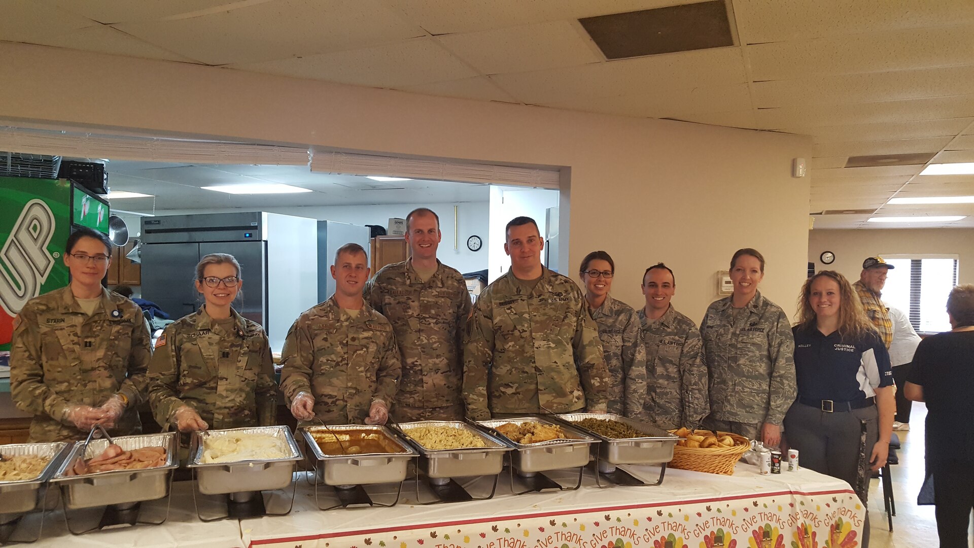 Eight uniformed military personnel and one mentee pose for a photo behind a food serving line