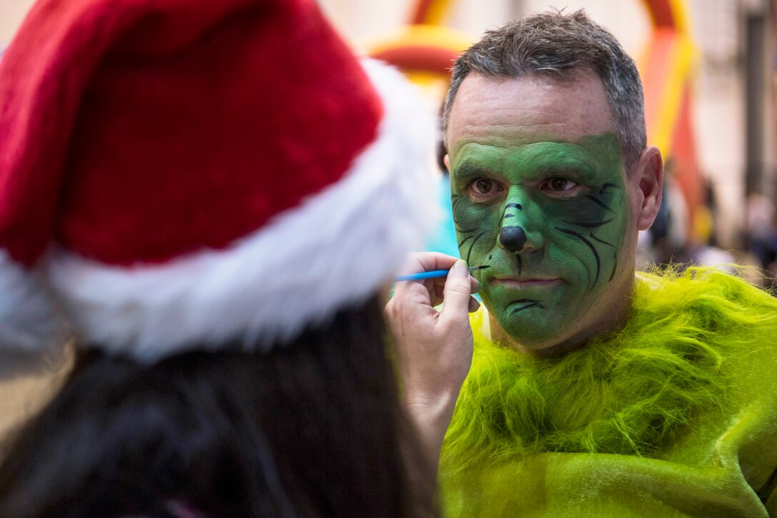 An airman in a green costume has his face painted green by someone in a Santa cap.