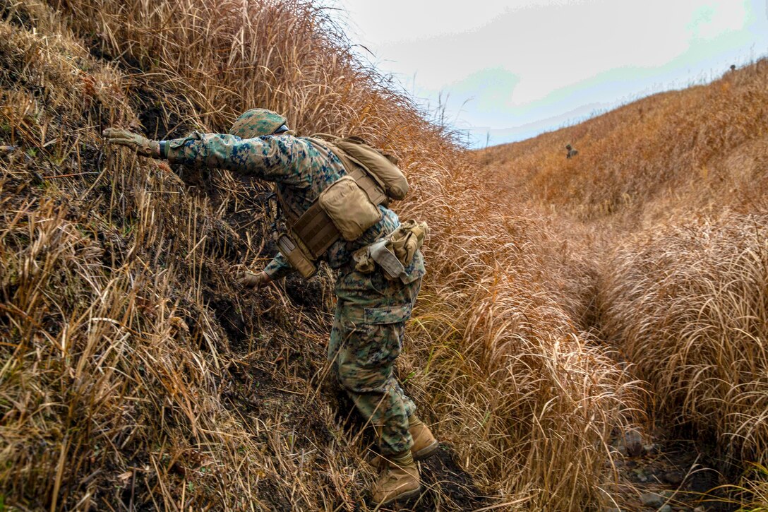 A Marine climbs a tall hill covered in grass.