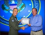 Photo of a male in a blue shirt and khaki pants receiving a certificate from another man in a Navy flight suit