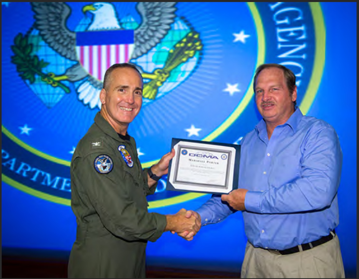 Photo of a male in a blue shirt and khaki pants receiving a certificate from another man in a Navy flight suit