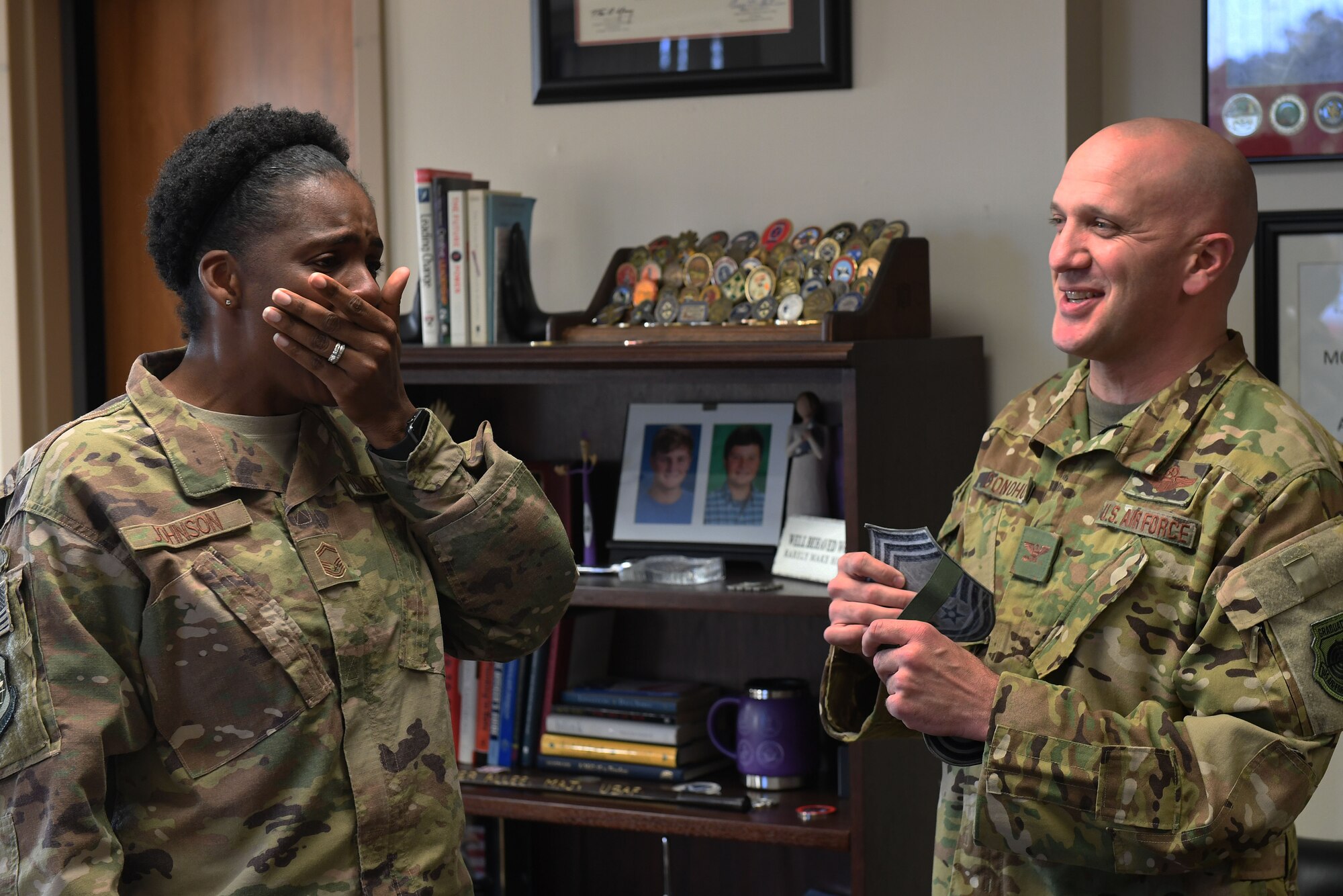 A black woman in camouflage uniform covers her mouth in surprise when a bald white man hands her new rank.