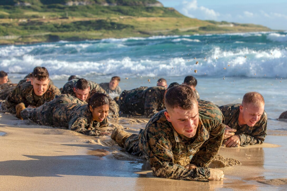 Marines crawl on the beach sand along the water.