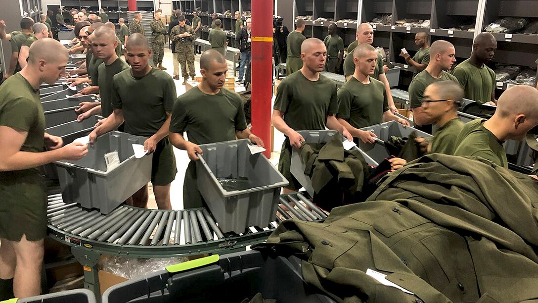A room full of young men at boot camp is gathered along a conveyor belt with bins to receive dress uniforms.