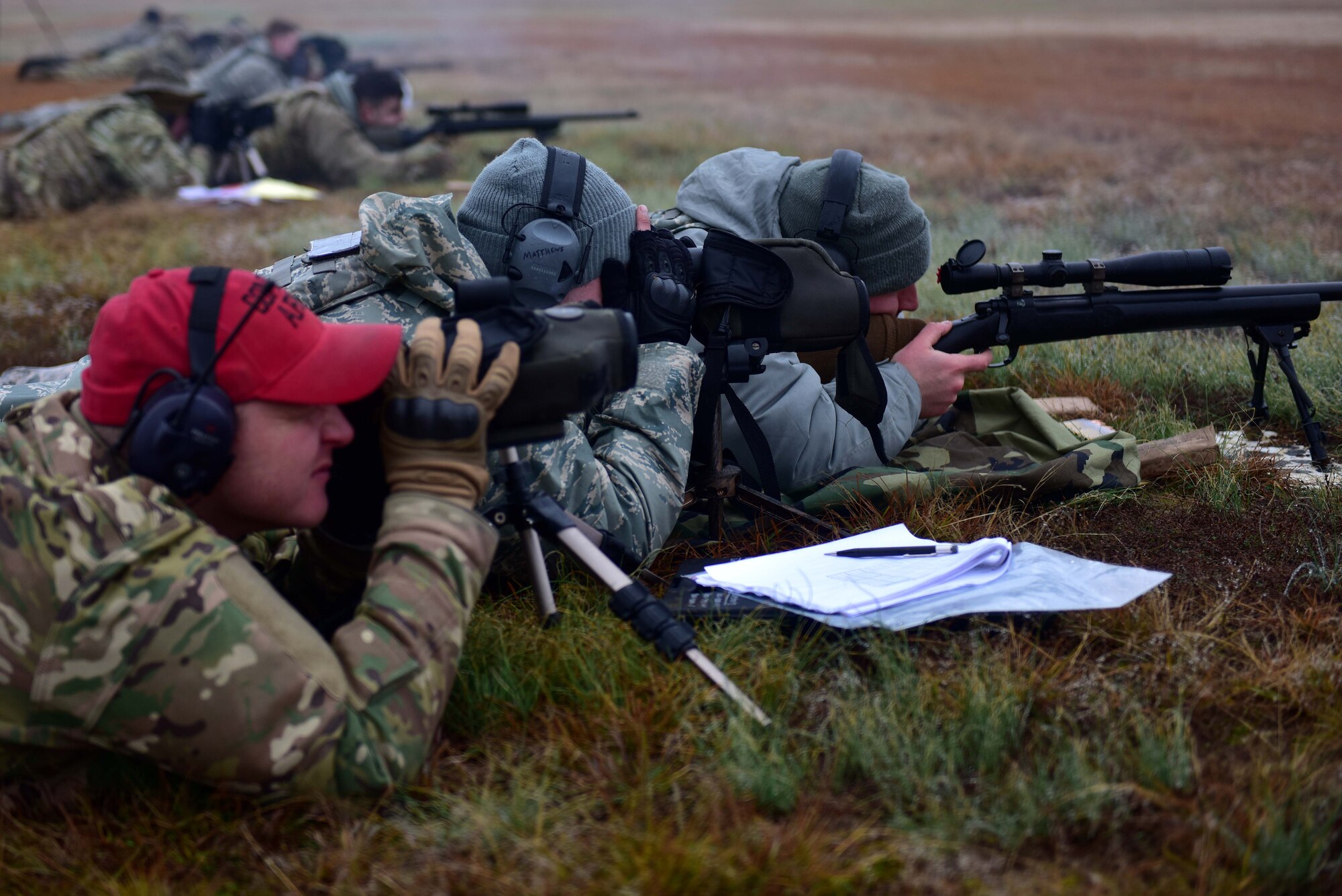 A man wearing  red hat and the operational camouflage pattern uniform looks down a scope at a range with two men wearing the Airman Battle Uniform.