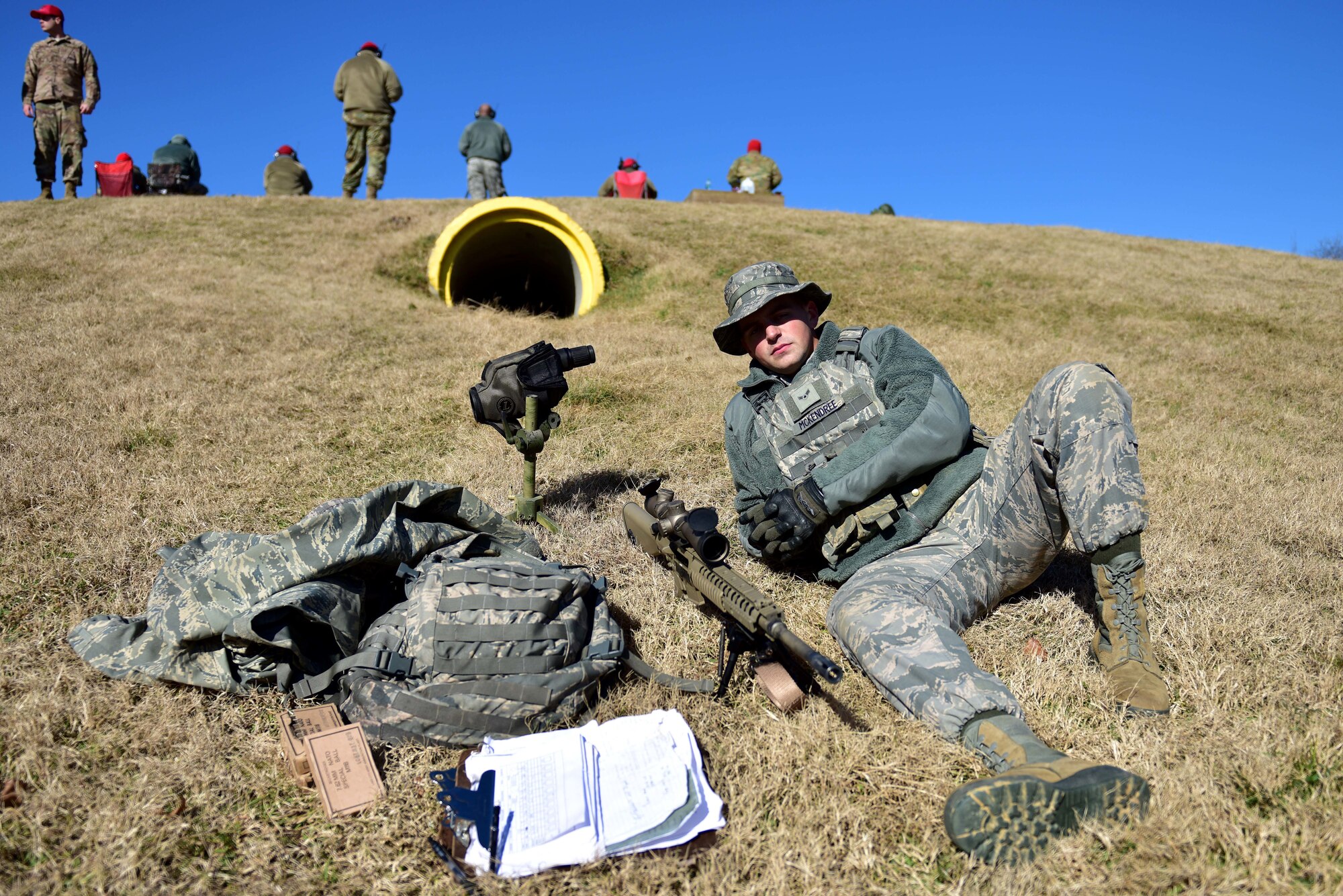 A man wearing the Airman Battle Uniform sits on a hill with a rifle, spotting scope, vest, backpack and clipboard.
