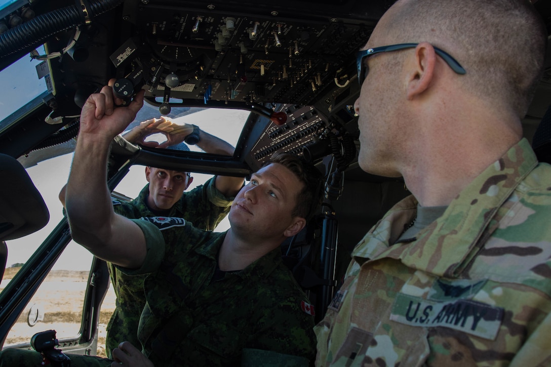 A soldier adjust controls in a helicopter cockpit as others watch.