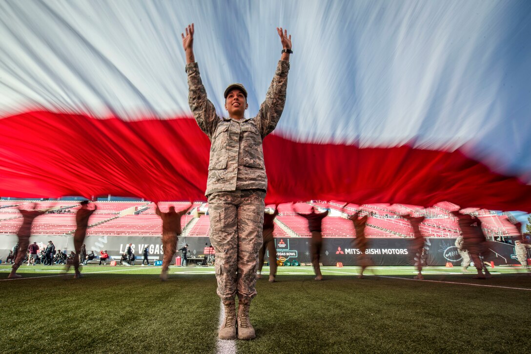 An airman holds her arms up on a football field, under giant red and white stripes of a U.S. flag.