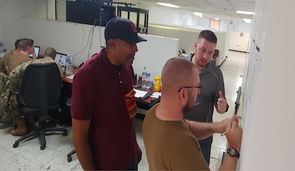 U.S. Navy Chief Timothy Bobrow, center, diagrams network architecture with Kenneth Simmons, J6, left, and Ben Loader, Department of Homeland Security Incident Response Manager, right, while Army and Navy personnel analyze network data in the back left of the room at Soto Cano Air Base, Dec. 2018.