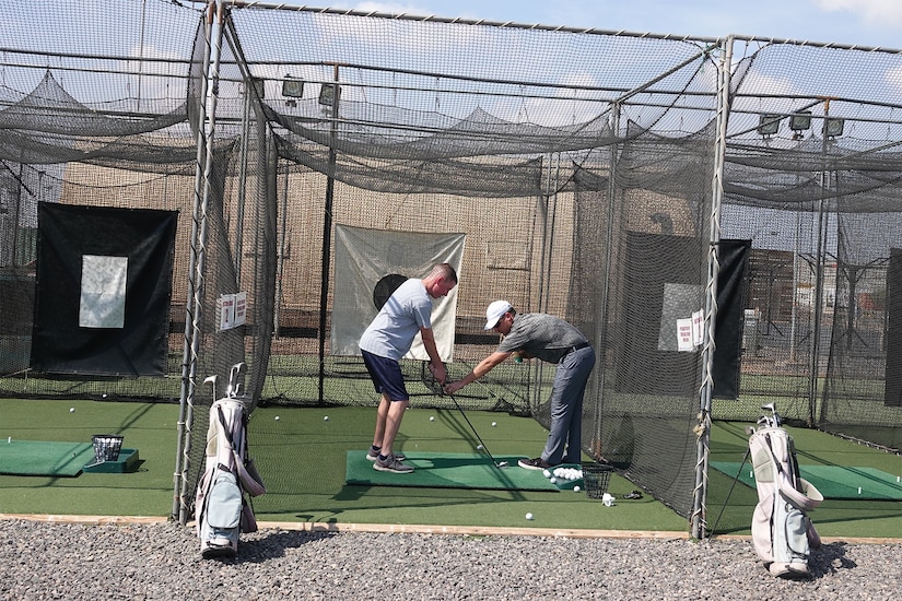 A golfer helps another man with his golf swing in a caged-in area.