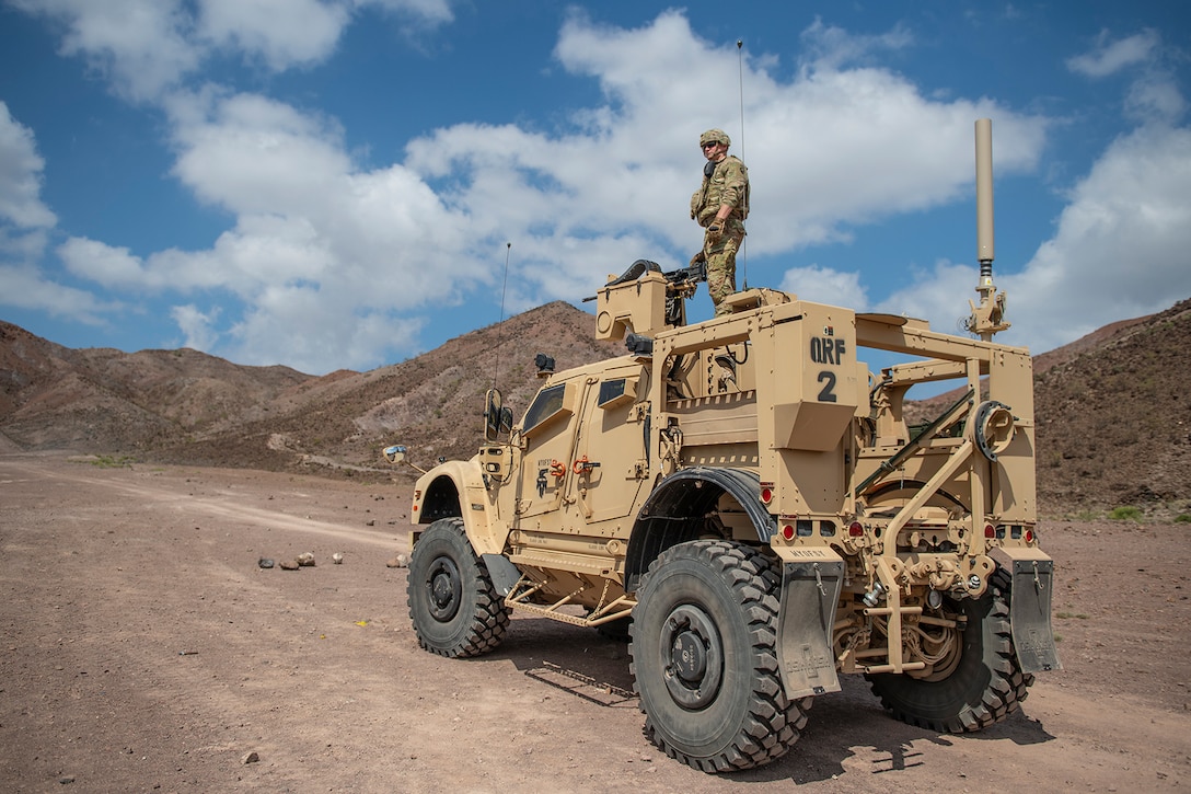 A service member stands on a vehicle while preparing a weapon on a military range.