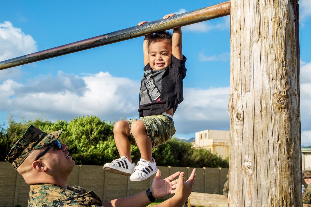 A service member stands under his toddler son, who hangs from a metal bar.