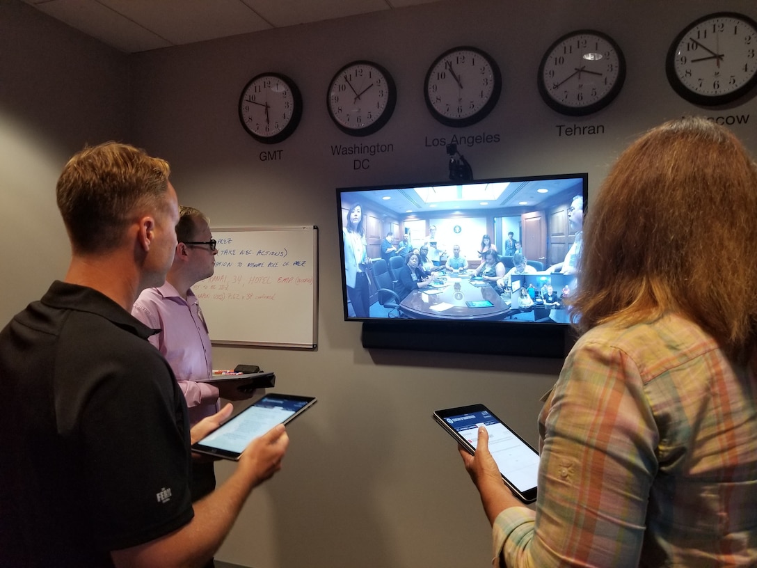 Three people stand in front of a television monitor during a teleconference